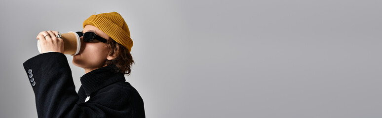 A young man in a black coat and yellow beanie sips coffee, showcasing a trendy vibe in a studio.