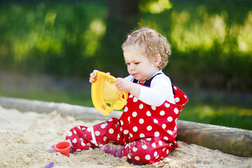 Cute toddler girl playing in sand on outdoor playground. Beautiful baby in red gum trousers having...