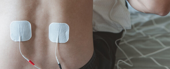 A person using electrical stimulation pads on their lower back for therapy or pain relief on a bed.