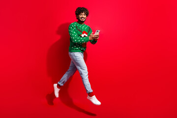 Full body photo of attractive young man hold device running jump dressed green christmas sweater theme party on red color background