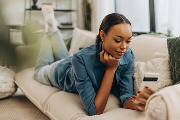 Obraz premium Smiling, African American woman holding mobile phone, social media lying on comfortable sofa