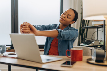 Portrait of tired, happy African American woman stretching hands up sitting at workplace