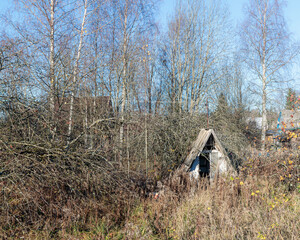 An old, decaying wooden structure surrounded by autumnal nature on an overgrown grassy lot,...