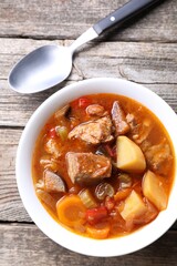 Delicious stew with vegetables in bowl and spoon on wooden table, top view