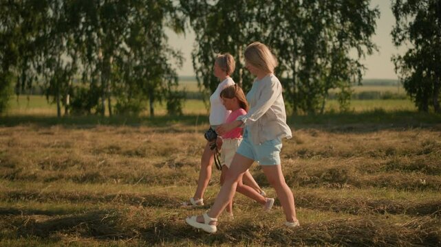 Child walks with her two aunts through vast open field, one aunt holding rope and pointing towards something in distance, while trees form a lush green background, all enjoying outdoor stroll