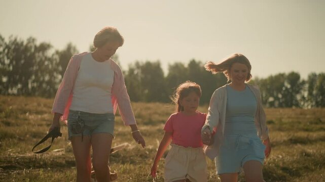 Child holding her mum's hand looks up as her mum smiles warmly, aunt walks alongside holding rope, gesturing for dog to come forward on sunny day in vast farmland with distant trees in background