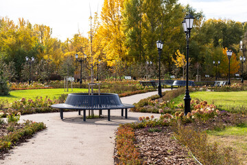 Round black metal bench in landscaped park, surrounded by flower beds and trees with golden autumn leaves, offering picturesque and tranquil atmosphere