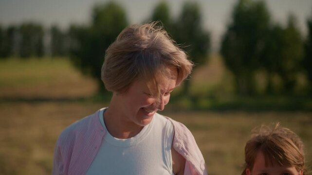 Close-up of little girl walking alongside aunt as aunt smiles warmly and touches her hair in breezy farmland setting, background features blurred trees and open field under sunlight