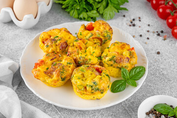Egg muffins or egg bites with bacon, cheese, vegetables and herbs on a white plate on a concrete...