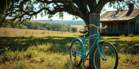 Fototapeta premium A turquoise bicycle leans against a weathered wooden post in a picturesque rural setting, its wheels framing a rustic farmhouse and a sprawling green field, creating a serene and timeless scene.