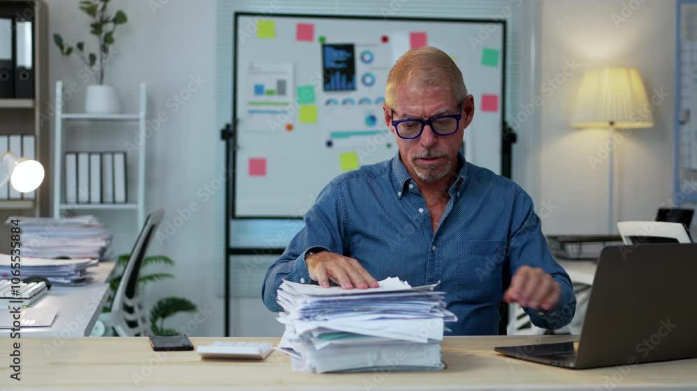 Poster accountant working on a laptop while managing a heavy workload of paperwork in a busy office environ