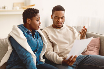 Obraz premium Financial Crisis Concept. Portrait Of Confused Perplexed African American Man Holding Paper Document And Reading Bank Notification Or Eviction Notice, Scared Woman Sitting On The Sofa In Living Room