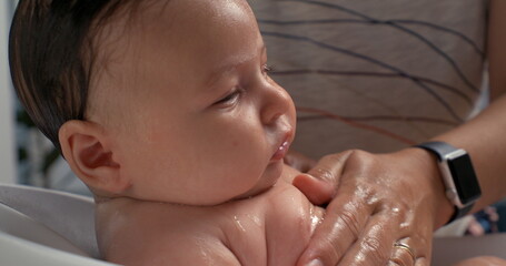 Hand gently supporting baby’s back during bath, baby’s face relaxed and calm, tender bathing...