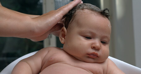 Hand gently washing baby’s head with water during bath, baby calm and relaxed, water droplets...