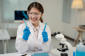 Fototapeta premium Young scientist wearing lab coat and gloves smiling while comparing two test tubes containing blue liquid, working in modern laboratory with microscope and computer