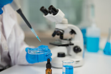 Close-up of scientist wearing gloves dropping blue liquid with micropipette on petri dish next to...