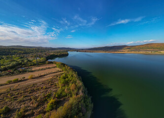 Panorama with a storage dam viewed from a drone in the countryside. A calm river that comes from the mountains among the green forests and flows into the lake at the edge of a village in Romania