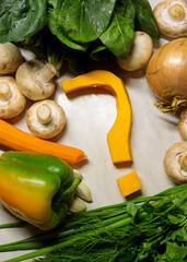 A question mark shaped from orange pumpkin lies among fresh vegetables: mushrooms, bell pepper, spinach, onion, garlic, carrot, and herbs on a white surface. A creative culinary arrangement