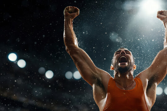 A wrestler exuberantly raises his arms in victory after winning a tough match, illuminated by bright lights in a packed stadium. The atmosphere is electric and intense