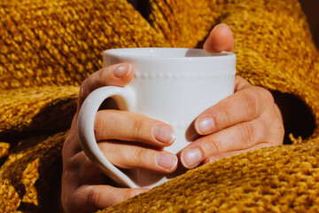 White cup in female hands. Woman wrapped in yellow plaid is drinking hot- tea or coffee. Good...