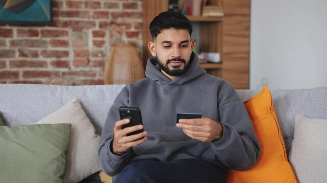 Concentrated male looking at credit card and cellphone while sitting on comfortable furniture indoors. Young customer of online banking service entering details for registration in account.