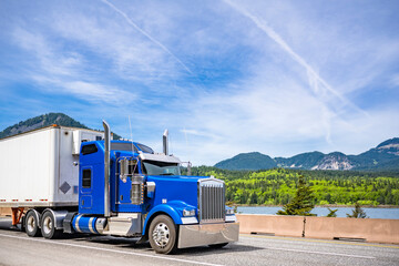 Bright blue classic big rig semi truck tractor with chrome parts transporting cargo in refrigerated...