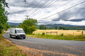Compact white cargo mini van making local delivery running on the winding road with summer meadow...