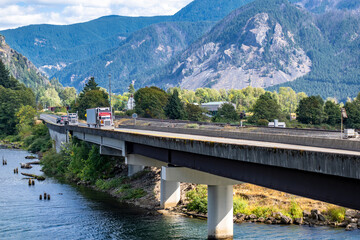 Fototapeta premium Red big rig classic semi truck transporting cargo in refrigerator semi trailer running in front of another semi trucks on the bridge across the tributary of the Columbia river in Columbia Gorge area