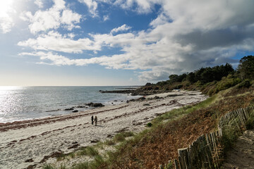 Deux promeneurs avancent le long de la plage de Beg Meil, entourés de rochers et de sable, sous un ciel bleu ponctué de nuages blancs d'automne.