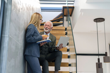 A senior Caucasian man in business attire discusses documents with a mid adult woman on a laptop, exemplifying teamwork in a sleek office setting.