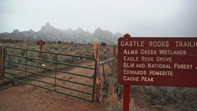 A large red trail sign and gate leading into Castle Rocks State Park, Idaho with a snowstorm in the distance