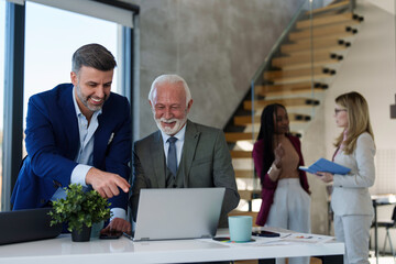 Close up of two Caucasian businessmen, one mature and one middle-aged, focused on a task on their laptop in a modern office setting with colleagues in the background.