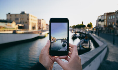Capturing a beautiful moment in Aveiro, Portugal, as hands hold a smartphone to take a photo of colorful boats and houses along the canal during sunset. Showcasing vibrant cityscape and architecture