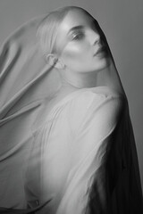A black-and-white portrait of a young woman behind a sheer fabric that gently frames her face,...