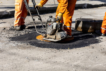 Workers repairing a road surface with asphalt in an urban area on a sunny day using a plate...
