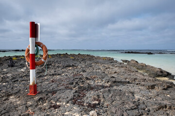 a pole with a lifebuoy in a rocky cove, on a day with overcast sky, lonely volcanic rock beach on a...