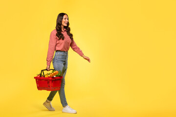 Full body length of young woman with red basket full of food products walking on yellow studio background. Shopping in supermarket concept © Home-stock