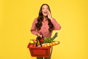 Young excited woman holding shopping basket full of food shouting with mouth wide open, announcing...