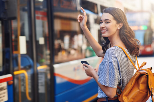 Girl, phone and happy at bus stop in city with hand, sign and backpack for travel to university. Person, smartphone and college student with smile, transport or commute on road for education in Spain - Powered by Adobe