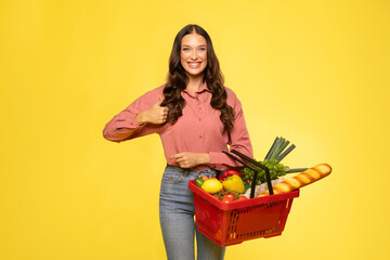 Cheerful European woman holding shopping basket full of food and giving thumbs up gesture, recommend supermarket, posing over yellow background