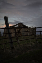 old abandoned dovecote with a partially collapsed roof, set against a dramatic evening sky. The surrounding grass and distant buildings add a rural charm, highlighting the beauty of decay and solitude