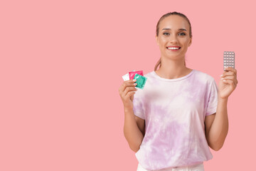 Young woman with condoms and contraceptive pills on pink background