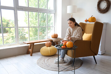 Beautiful young happy woman with pumpkins sitting on armchair in room decorated for autumn