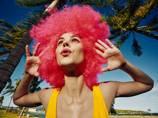 Pink wigged woman smiling, standing in front of a palm tree with hands on her head, bright day,...