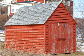 The exterior of a small vintage red wooden tool shed with faded Cape Cod clapboard siding, double wood doors, and a cedar-shingled peaked roof. The fishing storage hut is in a grassy farm backyard.