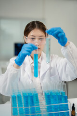 Young scientist wearing lab coat and gloves comparing two test tubes with blue liquid while working...