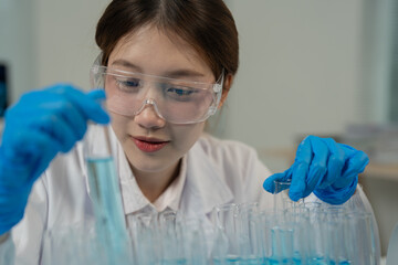Young female scientist wearing protective glasses and gloves carefully examining test tubes filled...