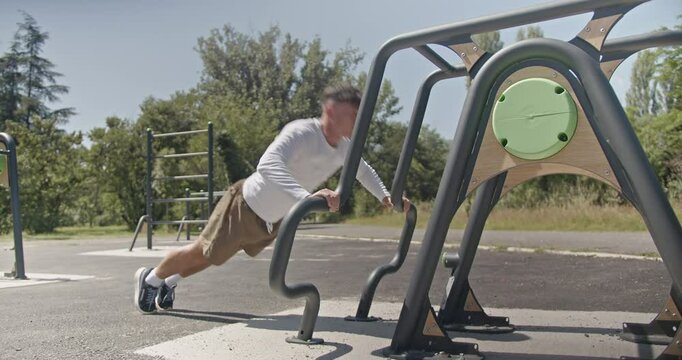 A fit man doing push ups on parallel bars in a park, on a sunny day. He completes his set and gets up.
