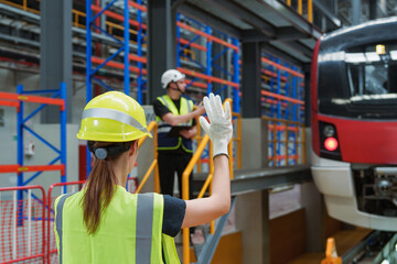 Female traffic controller in safety vest jacket with green hard hat gives signal to the driver of electric locomotives red train in the high tech maintenance plant.