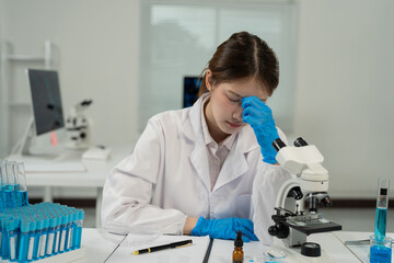 Young scientist wearing lab coat and gloves is having a headache while working with microscope and...